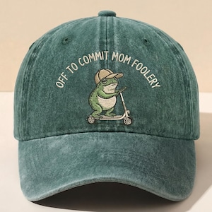 May include: Teal baseball cap with the embroidered phrase "OFF TO COMMIT MOM FOOLERY." The cap features a cartoon frog wearing a hat and riding a scooter. The cap is made of a textured fabric.