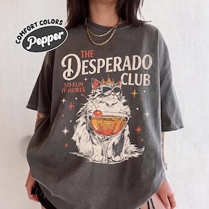 Puede incluir: Una camiseta gris carbón de manga corta con el gráfico de un gato con corona y gafas de sol, sosteniendo una copa de cóctel con una cereza. El texto dice THE DESPERADO CLUB y SO FUN IT HURTS. Un logo en la esquina dice COMFORT COLORS Pepper.