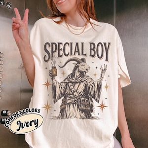 Puede incluir: Una camiseta de color marfil con el gráfico de una cabra con túnica que sostiene un frasco humeante y el texto SPECIAL BOY. El diseño incluye estrellas y una alforja. La camiseta tiene cuello redondo y mangas cortas con etiqueta visible.