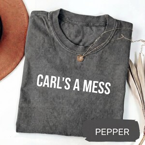 May include: A dark gray t-shirt with the text "CARL'S A MESS" in white. A silver necklace with a small pendant is draped across the shirt. The word "PEPPER" is in a dark gray rectangle at the bottom of the shirt.