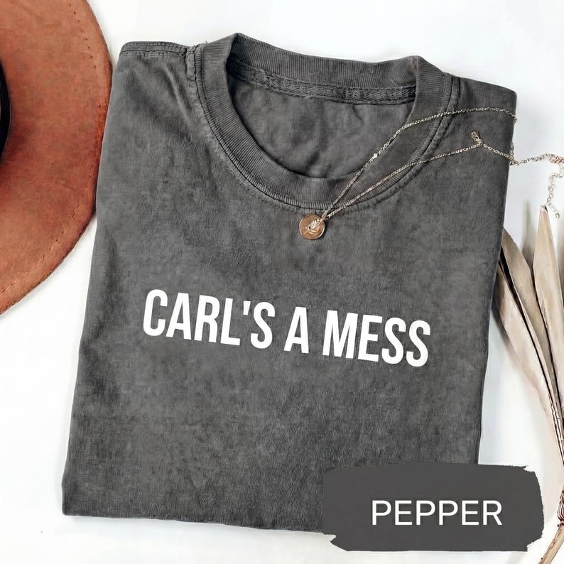 May include: A dark gray t-shirt with the text "CARL'S A MESS" in white. A silver necklace with a small pendant is draped across the shirt. The word "PEPPER" is in a dark gray rectangle at the bottom of the shirt.