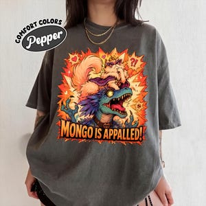 May include: A charcoal gray t-shirt featuring a graphic print of a fluffy cat wearing a crown and cape sitting on a blue dinosaur. The text reads "COMFORT COLORS Pepper" and "MONGO IS APPALLED!" with an orange and yellow explosive background.