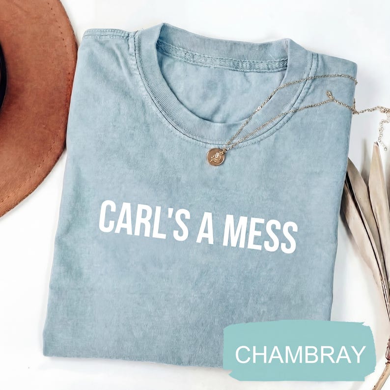 May include: A light blue chambray t-shirt with the text "CARL'S A MESS" in white block letters. A gold-colored necklace with a pendant is draped on the shirt. A brown hat is partially visible in the upper left corner.