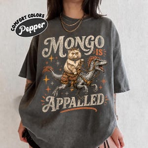 May include: A charcoal gray short-sleeve t-shirt featuring a graphic of a fluffy cat riding a dinosaur. The text reads "MONGO IS APPALLED" in a distressed font. The design includes orange star accents and a saddle on the dinosaur's back.