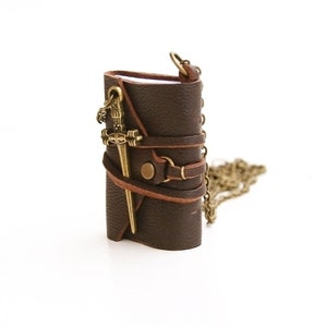 May include: A brown leather journal with a metal sword charm and a brown leather strap. The journal is attached to a chain.