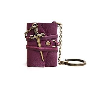 May include: A small purple leather notebook with a metal sword charm attached to a chain. The notebook is wrapped with leather straps and has a metal button closure.
