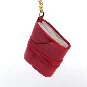 May include: A miniature book pendant featuring a bright red textured leather cover wrapped with a matching red leather cord. The small book has visible white pages and is attached to a thin gold-toned metal chain by a small jump ring.