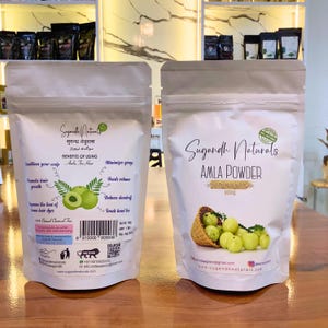 May include: Two white pouches of Sugandh Naturals Amla Powder. One pouch has text in multiple languages, including "Benefits of Using Amla For Hair." The other pouch shows a basket of green amla fruit and the text "AMLA POWDER 100g."