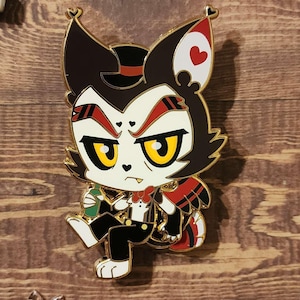May include: A black and white enamel pin of a cartoon cat wearing a tuxedo and a top hat. The cat has red eyes and a red heart on its ear. The pin is on a wooden background with a bottle of wine and playing cards.