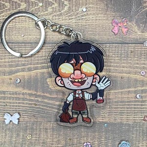 May include: A clear acrylic keyring featuring a cartoon character with black hair, large yellow goggles, and a brown apron. The character is waving and holding a bag and a vial. The keyring has a silver ring and chain.