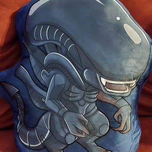 May include: A plush pillow in the shape of an Alien Xenomorph. The pillow is primarily dark blue with a detailed illustration of the creature in shades of gray and white. The pillow is soft and plush, perfect for fans of the Alien franchise.