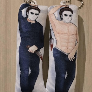 May include: Two rectangular plush keychains featuring a stylized illustration of a character in a mask. One keychain depicts the character in a dark blue jumpsuit holding a bloody knife. The other shows the character shirtless in blue jeans.