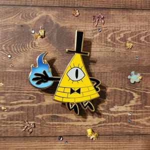 May include: A gold-colored enamel pin featuring Bill Cipher, a triangular character with a single eye, a black bow tie, and a black top hat. Bill Cipher is holding a blue flame in his right hand.