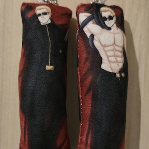 May include: Two rectangular plush keychains. One features a man in a black coat and sunglasses, the other a shirtless man with sunglasses. Both have red and black backgrounds and silver keychains.