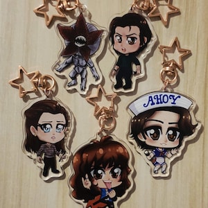 Puede incluir: Una colección de llaveros de acrílico transparente con personajes de dibujos animados. Cada llavero tiene un cierre en forma de estrella de color dorado. Los personajes incluyen un Demogorgon, una persona con gorro de marinero con la palabra "AHoy" y otros personajes.