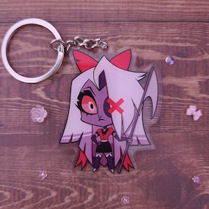 May include: A clear acrylic keychain featuring a cartoon character with purple skin, red hair, and a red bow. The character is holding a scythe and has a red X over one eye.