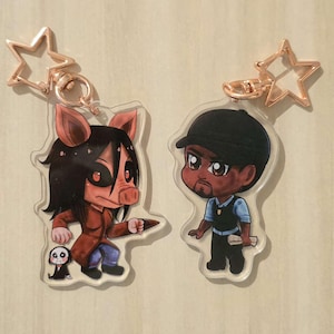 Könnte beinhalten: Zwei Schlüsselanhänger aus Acryl mit Cartoon-Charakteren. Einer zeigt einen Charakter mit Schweinemaske, schwarzen Haaren und einem roten Mantel. Der andere zeigt einen Mann mit schwarzer Kappe und Weste. Beide haben sternförmige Verschlüsse.