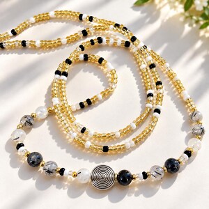 May include: A beaded necklace featuring translucent gold, opaque white, and black seed beads arranged in a repeating pattern. The strand includes larger clear beads with black inclusions, solid black spheres, and a central silver spiral charm.
