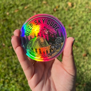 May include: A holographic sticker with a rainbow iridescent finish. The sticker features a cartoon illustration of a campsite with a tent, a person looking through a telescope, and a shooting star. The text "Deep Sky Dude" is written in a circle around the illustration. The text "Dark Skies Are Calling And I Must Go" is written in a smaller circle around the bottom of the sticker.
