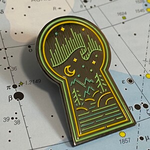 Puede incluir: Un pin de esmalte con forma de ojo de cerradura con un diseño verde, amarillo y negro. El diseño presenta un cielo nocturno con estrellas, una luna creciente y una cordillera. El pin es una excelente manera de agregar un toque de fantasía a tu mochila, chaqueta o bolso.