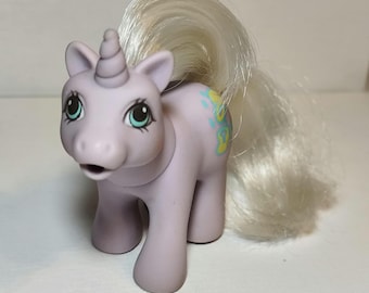 May include: A light purple unicorn toy with a white mane and tail. The unicorn has large teal eyes, a spiraled horn on its forehead, and a yellow and blue graphic design on its flank. The figure is standing in a forward-facing position.