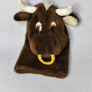 Puede incluir: Títere de toro de peluche marrón con cuernos, orejas blancas y un anillo nasal amarillo. El títere tiene ojos grandes y redondos con pupilas negras y reflejos anaranjados. Un mechón de pelo blanco es visible en la parte superior de la cabeza.