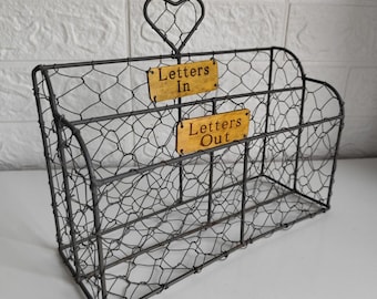 May include: A dark grey wire mesh mail organiser featuring two compartments with yellow rectangular labels reading Letters In and Letters Out. The structure includes a decorative heart-shaped wire handle at the top centre.