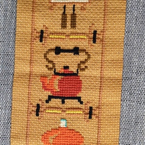 Könnte beinhalten: Ein brauner rechteckiger Wandbehang mit Kreuzstich und einem geometrischen Muster mit einer roten Teekanne, einer blau-weißen Lampe und einer gelben und orangefarbenen Blume. Das Muster ist mit einem braunen Rahmen eingefasst.