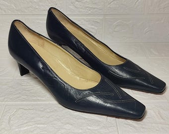 May include: A pair of navy blue leather court shoes featuring a square toe, a low kitten heel, and decorative tonal stitching on the vamp. The interior of the shoes is lined with a light beige material, providing a contrast to the dark exterior leather.