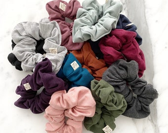 Könnte beinhalten: Eine Sammlung von 12 bunten Haargummis aus weichem Stoff. Die Haargummis sind in verschiedenen Farben, darunter Grau, Türkis, Burgunderrot, Rosa und Grün. Jedes Haargummi hat ein kleines weißes Etikett mit einem einfachen Design.