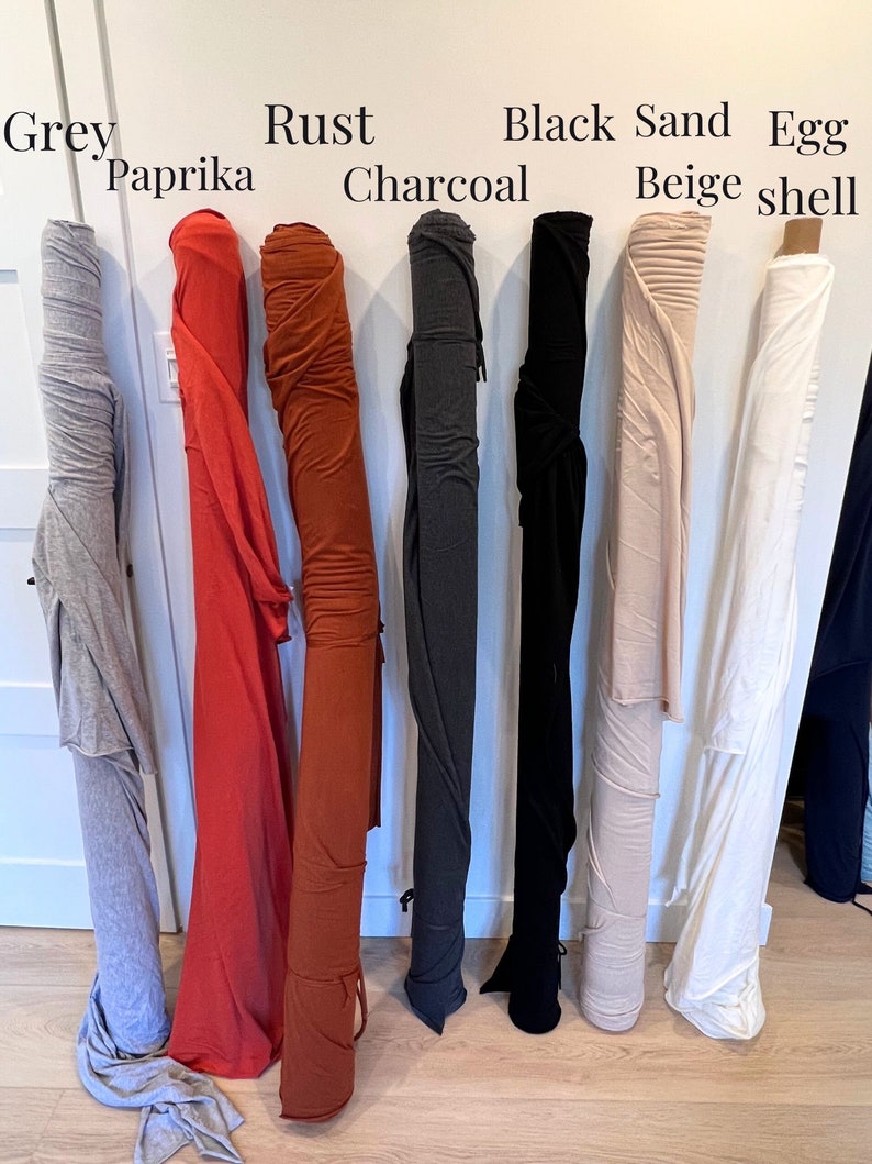 Puede incluir: Ocho rollos de tela en varios colores, incluyendo gris, paprika, &oacute;xido, carb&oacute;n, negro, beige arena y c&aacute;scara de huevo. La tela est&aacute; enrollada y colgando de una pared blanca.