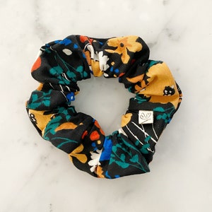 May include: A black scrunchie with a colorful floral print featuring yellow, orange, blue, and green flowers. The scrunchie has a small white tag with a leaf design.