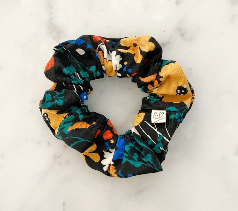 May include: A black scrunchie with a colorful floral print featuring yellow, orange, blue, and green flowers. The scrunchie has a small white tag with a leaf design.