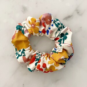 May include: A white scrunchie with a floral print featuring yellow, red, pink, blue, and green flowers. The scrunchie has a small white tag with a heart symbol.