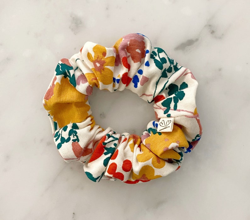 May include: A white scrunchie with a floral print featuring yellow, red, pink, blue, and green flowers. The scrunchie has a small white tag with a heart symbol.