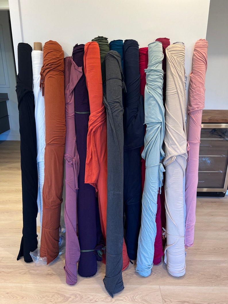 Puede incluir: Una colecci&oacute;n de rollos de tela en varios colores, incluyendo blanco, naranja, morado, rojo, gris, azul y rosa. Los rollos est&aacute;n apilados contra una pared blanca.