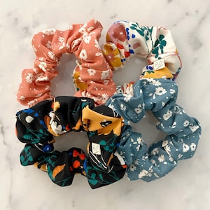 May include: Four floral patterned scrunchies in pink, blue, black, and white. The scrunchies are made of fabric and have a gathered design.