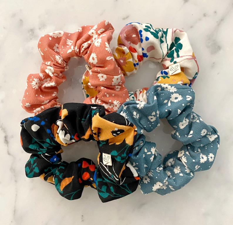 May include: Four floral patterned scrunchies in pink, blue, black, and white. The scrunchies are made of fabric and have a gathered design.