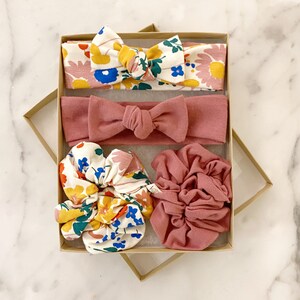 May include: A set of four hair accessories in a gift box. The set includes two headbands, one with a floral print and the other a solid pink color, and two scrunchies, one with a floral print and the other a solid pink color.