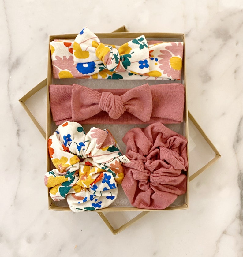 May include: A set of four hair accessories in a gift box. The set includes two headbands, one with a floral print and the other a solid pink color, and two scrunchies, one with a floral print and the other a solid pink color.