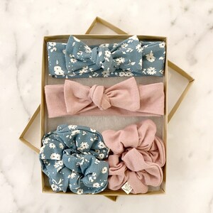 May include: A set of four hair accessories in a brown box. The set includes two headbands, one in a light blue floral print and one in a solid light pink. There are also two scrunchies, one in a light blue floral print and one in a solid light pink.