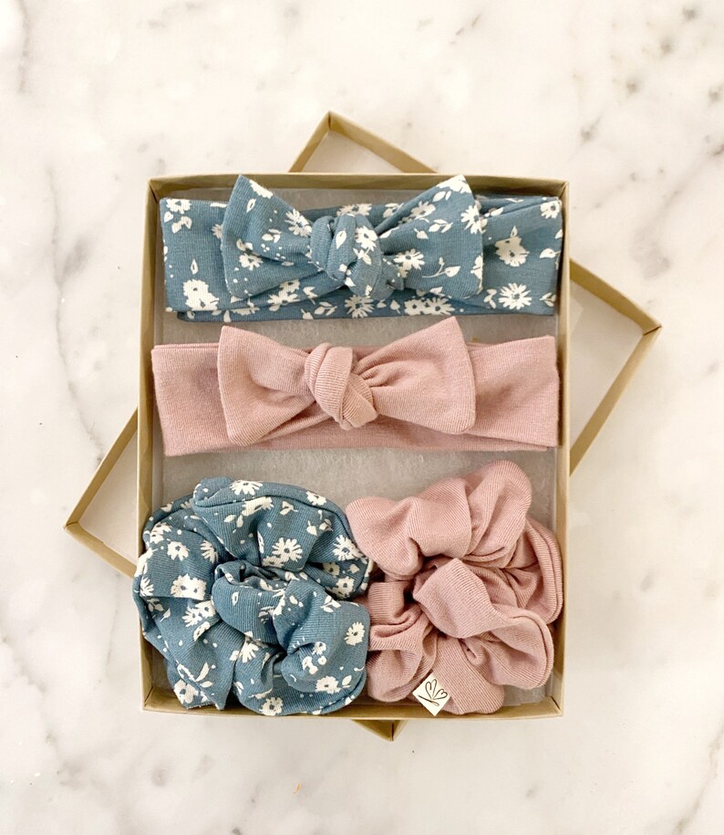 May include: A set of four hair accessories in a brown box. The set includes two headbands, one in a light blue floral print and one in a solid light pink. There are also two scrunchies, one in a light blue floral print and one in a solid light pink.