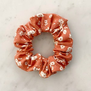 May include: A coral-colored scrunchie with a white floral pattern. The scrunchie is made of a soft, stretchy fabric and is perfect for holding hair in place.