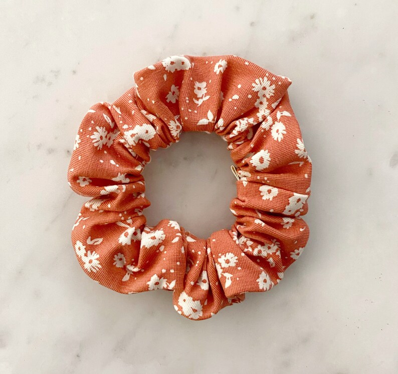 May include: A coral-colored scrunchie with a white floral pattern. The scrunchie is made of a soft, stretchy fabric and is perfect for holding hair in place.