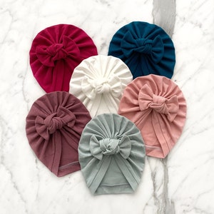 May include: Six different coloured turban headbands with a bow knot on top. The colours are burgundy, white, teal, pink, and two shades of brown.
