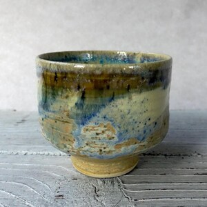 Peut inclure: Un bol en céramique fait à la main avec un glaçage unique. Le bol présente un mélange de couleurs, notamment le bleu, le vert et le beige, créant un aspect texturé et organique. Le bol a une forme arrondie avec une petite base circulaire.