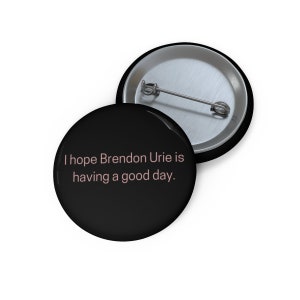 May include: Black button with a silver pin and the text "I hope Brendon Urie is having a good day."