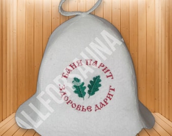May include: A white felt sauna hat with a hanging loop at the top. The front features an embroidered design of two green oak leaves centered within a circular arrangement of red Cyrillic text that reads Banya Parit Zdorovye Darit.