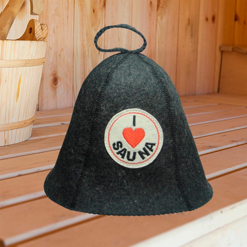 Puede incluir: Un gorro de sauna gris oscuro con un parche redondo que dice "I ❤️ SAUNA". El gorro tiene un lazo en la parte superior y est&aacute; hecho de un material grueso similar al fieltro. El fondo es una sauna de madera.