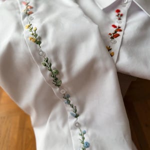 Può includere: Due camicie bianche con ricami floreali lungo la paramontura. Il ricamo presenta una varietà di colori, tra cui giallo, verde, blu e rosso. Le camicie sono realizzate in un tessuto liscio e croccante.